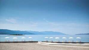 A beach with white umbrellas and lounge chairs, a small island, and mountains in the background.