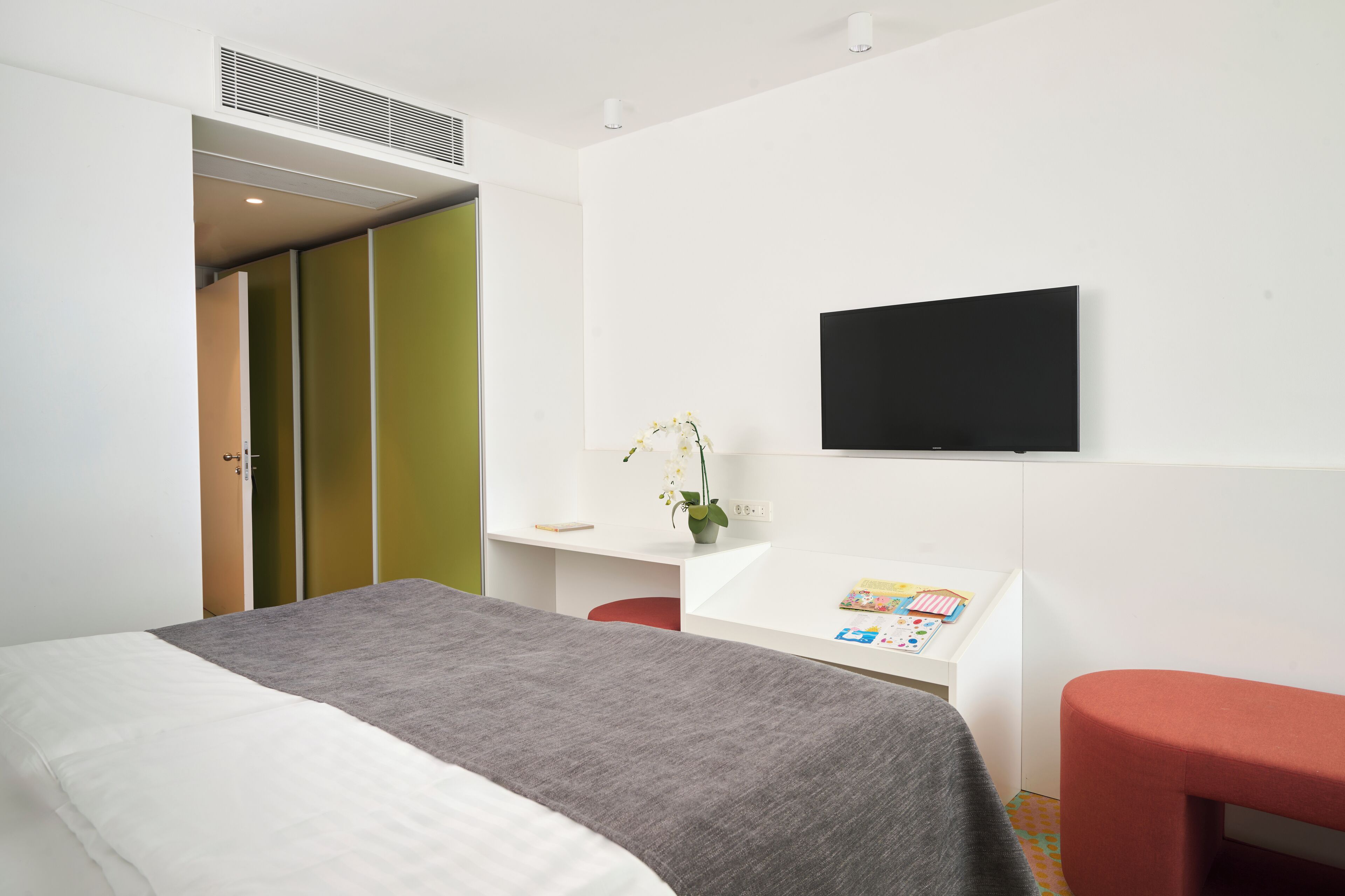 A modern hotel room with a bed, a desk with a flat-screen TV, a small table with a vase of flowers, and a red bench.