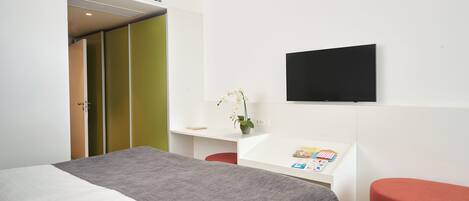 A modern hotel room with a bed, a desk with a flat-screen TV, a small table with a vase of flowers, and a red bench.