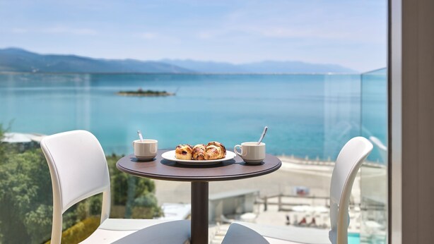 A table with a plate of pastries and two cups, overlooking a clear blue sea and distant mountains.