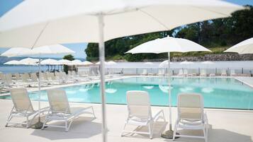Un espace aménagé au bord de la piscine, avec des chaises longues blanches et des parasols, une piscine à l’eau claire et un mur de pierre en arrière-plan.
