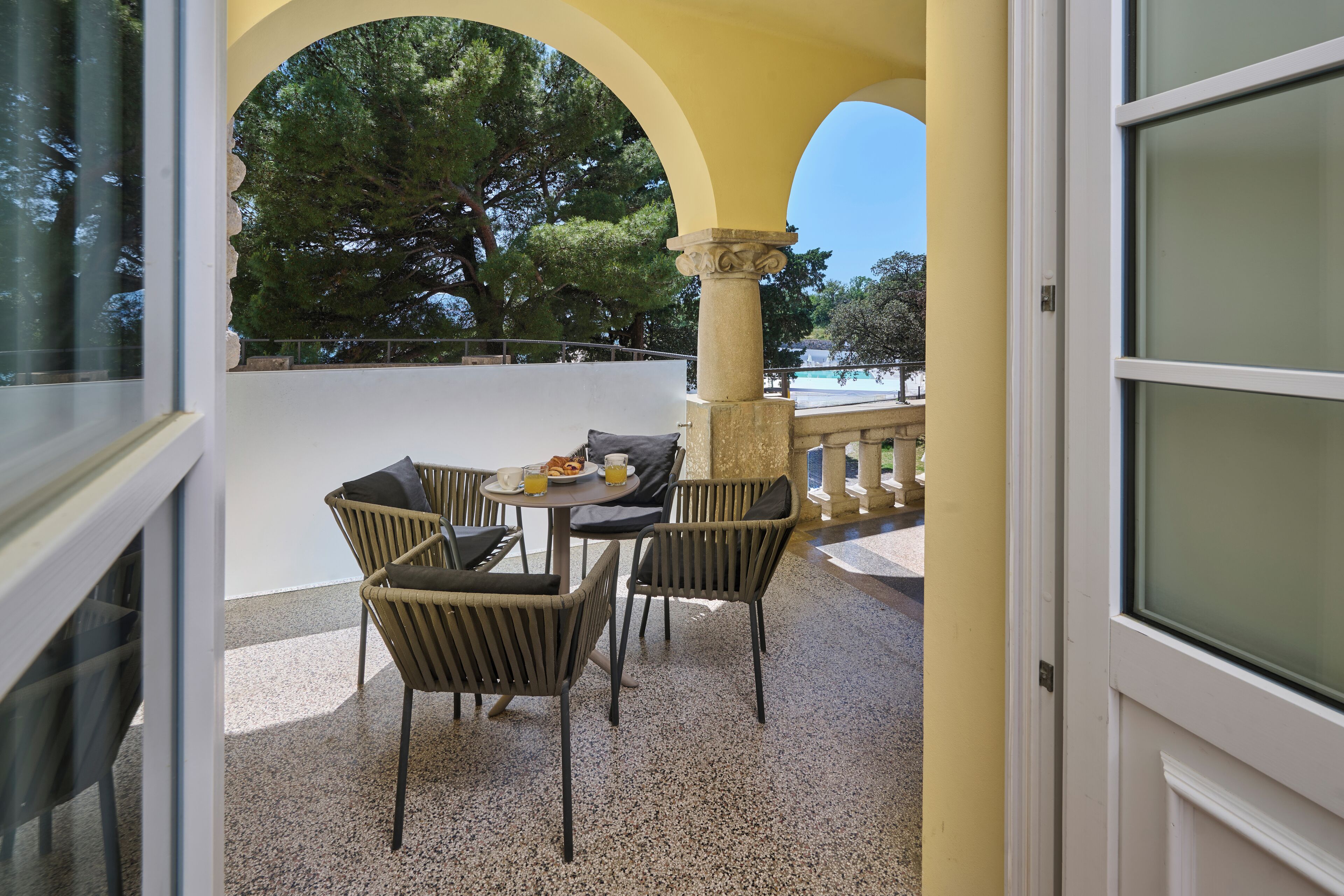 A balcony with a table and chairs, a view of trees, and a pool in the background.