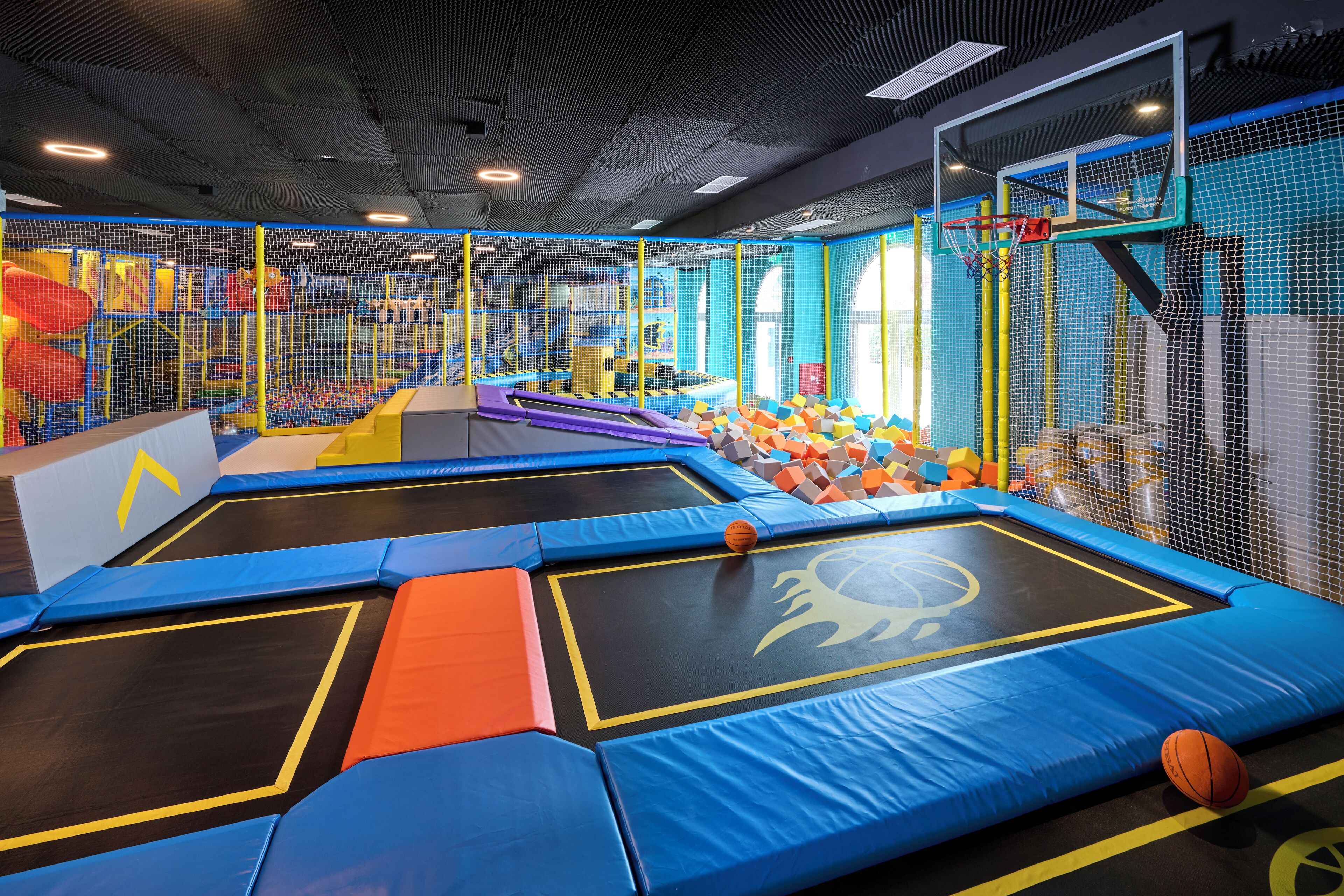 An indoor trampoline area with safety netting, padded flooring, and a basketball hoop.