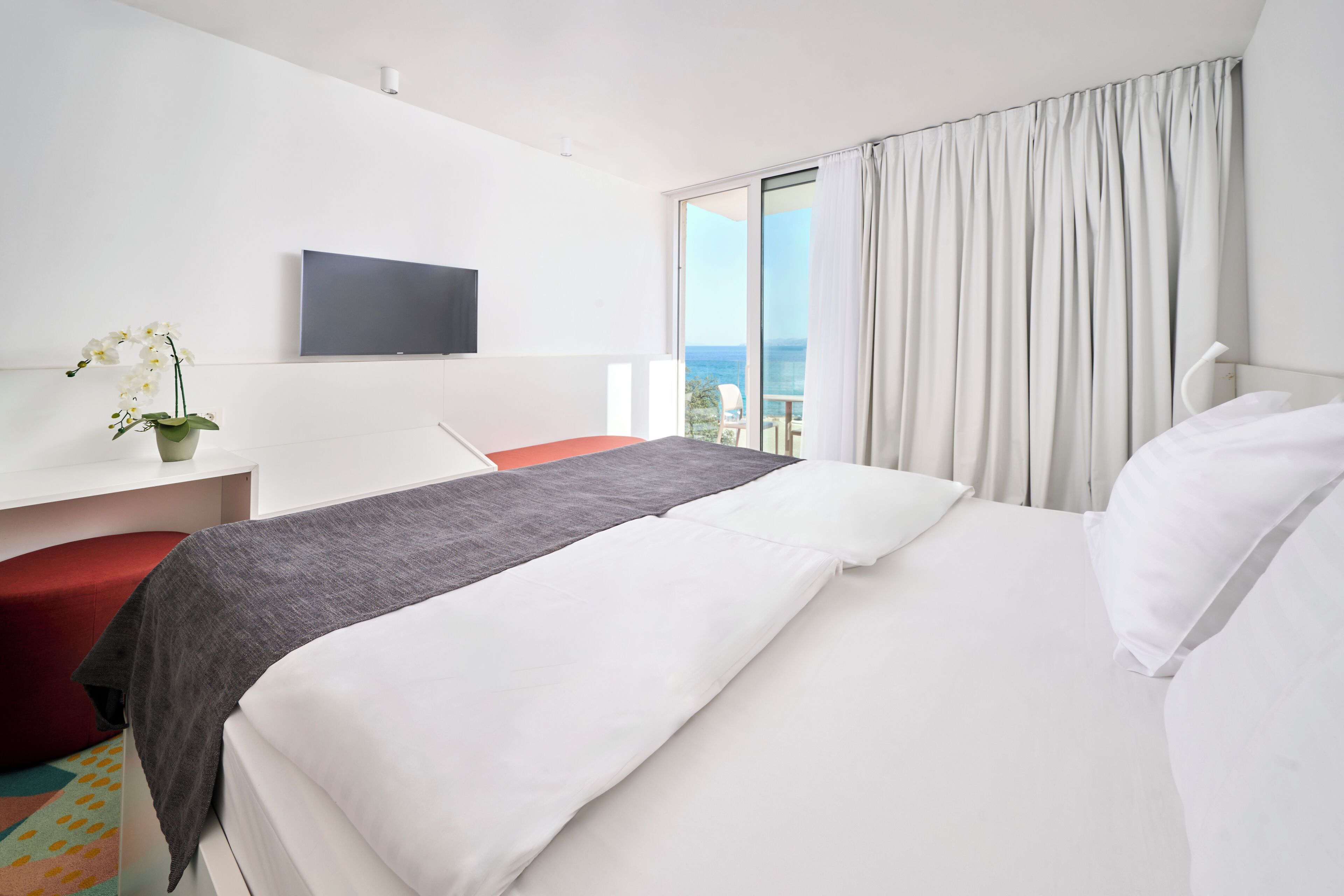 Une chambre d’hôtel avec un lit, un téléviseur fixé au mur, une fenêtre avec des rideaux et une vue sur l’océan.