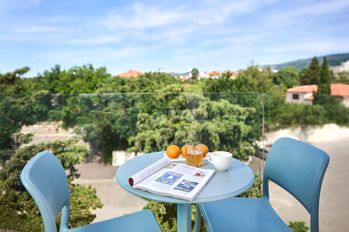 A round table with a newspaper, a glass of iced tea, and two oranges on a balcony overlooking a scenic view of trees and houses.