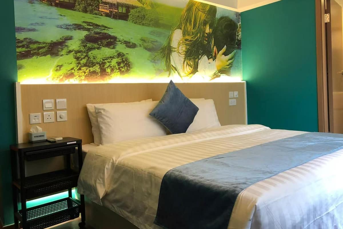 Ein Hotelzimmer mit einem Bett, einem Nachttisch, einer Wandleuchte und einem Wandgemälde mit tropischer Szenerie.