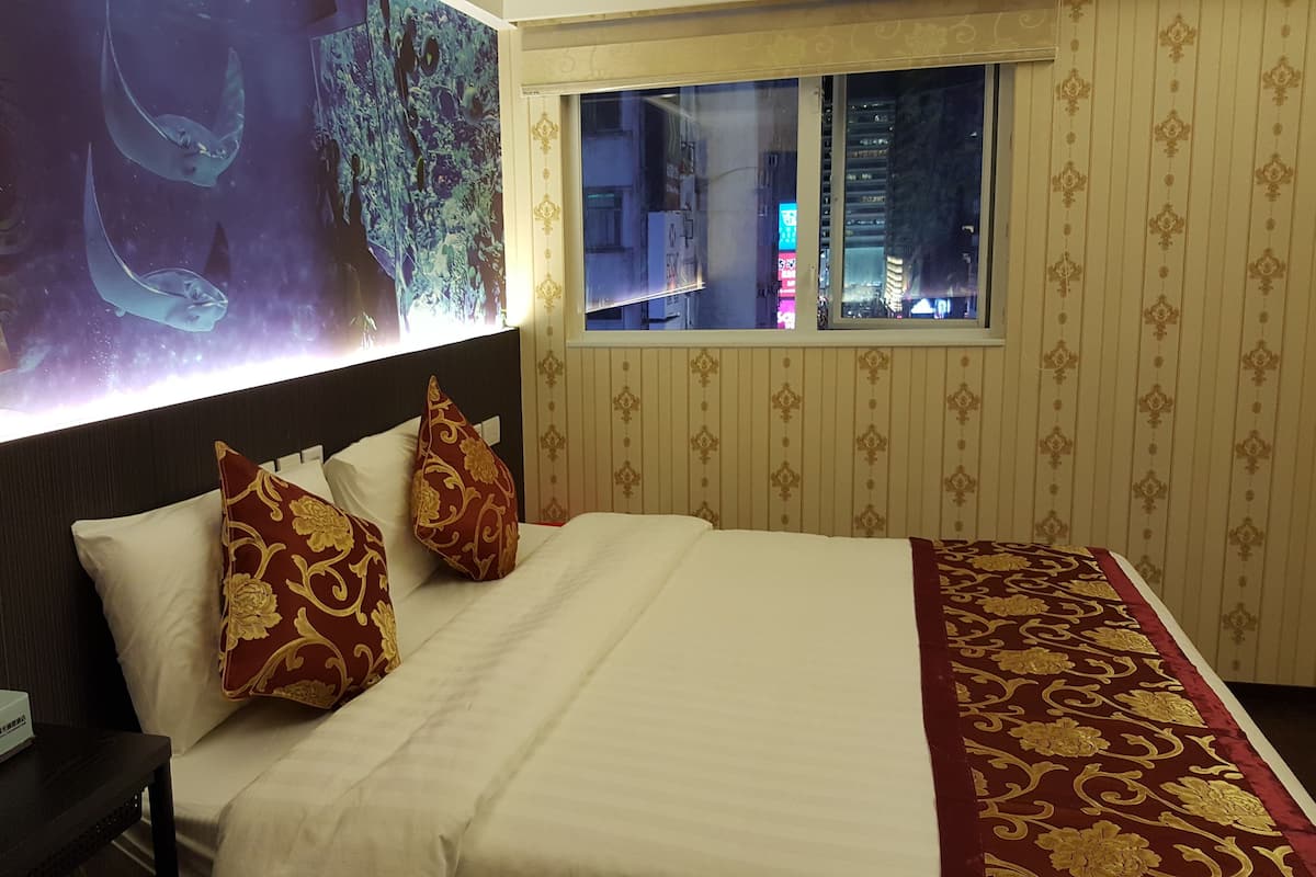 Ein Hotelzimmer mit einem großen Bett, zwei Zierkissen, einem Nachttisch mit Telefon und Taschentuchbox, einem Fenster mit Blick auf die Stadt und einer Wand mit einem großen abstrakten Gemälde.
