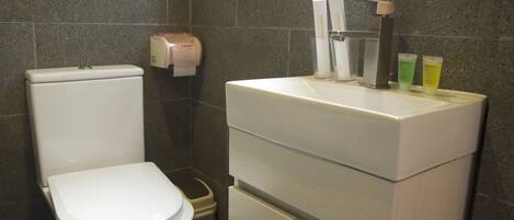 A bathroom with a toilet, a trash bin, a paper towel dispenser, and a sink with hand soap and lotion dispensers.