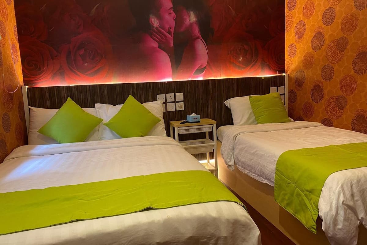 Ein Hotelzimmer mit zwei Betten, jeweils mit weißer Bettwäsche und grünen Kissen. Zwischen den Betten steht ein Nachttisch mit Taschentuchbox. Das Zimmer hat eine gemusterte Tapete und ein großes Blumenbild an der Wand.