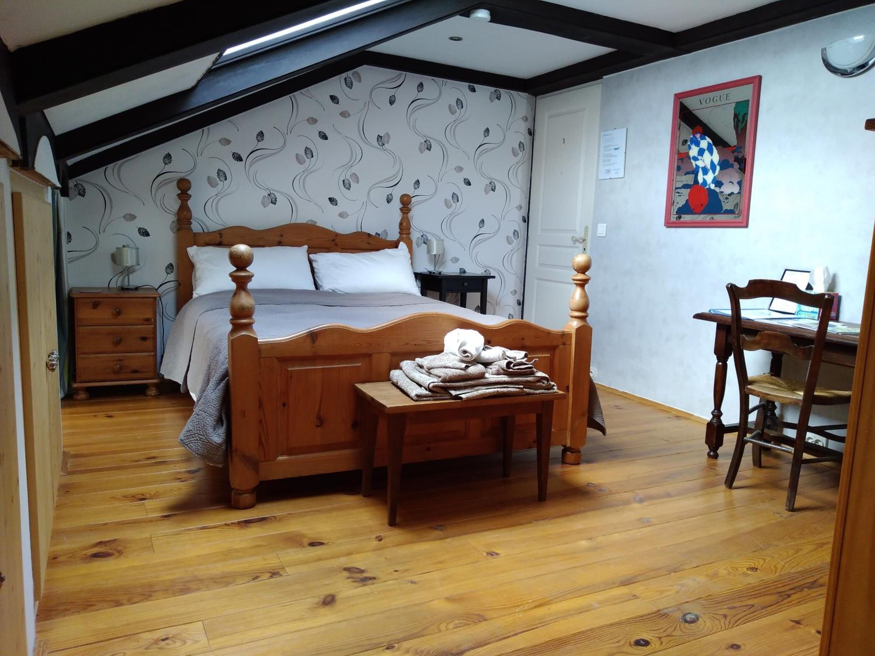 A bedroom with a wooden four-poster bed, a small bedside table with towels, a desk with a chair, and a wall-mounted mirror.