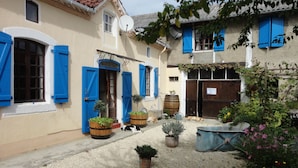 Un patio con un edificio que tiene contraventanas azules, plantas en macetas y un barril.