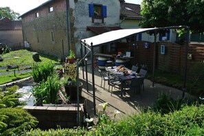 En terrasse med bord og stole, en lille have med planter og et bassin, samt en bygning med et tegltag i baggrunden.