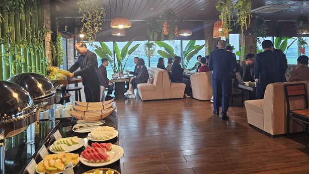 Zona de bufé con diferentes opciones de comida y una persona sirviendo. Hay mesas y sillas dispuestas para los huéspedes, y la sala tiene un diseño moderno con plantas colgantes e iluminación colgante.