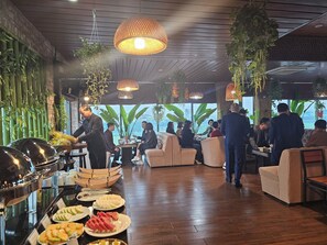 Een buffetgedeelte met diverse gerechten en een persoon die bedient. Er staan tafels en stoelen klaar voor gasten, en de ruimte heeft een modern design met hangplanten en hanglampen.