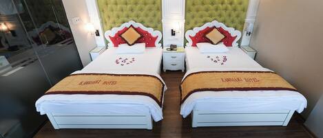 A hotel room with two beds, each with a headboard and a pillow with a heart-shaped design. The room has a green upholstered headboard and a white wall.