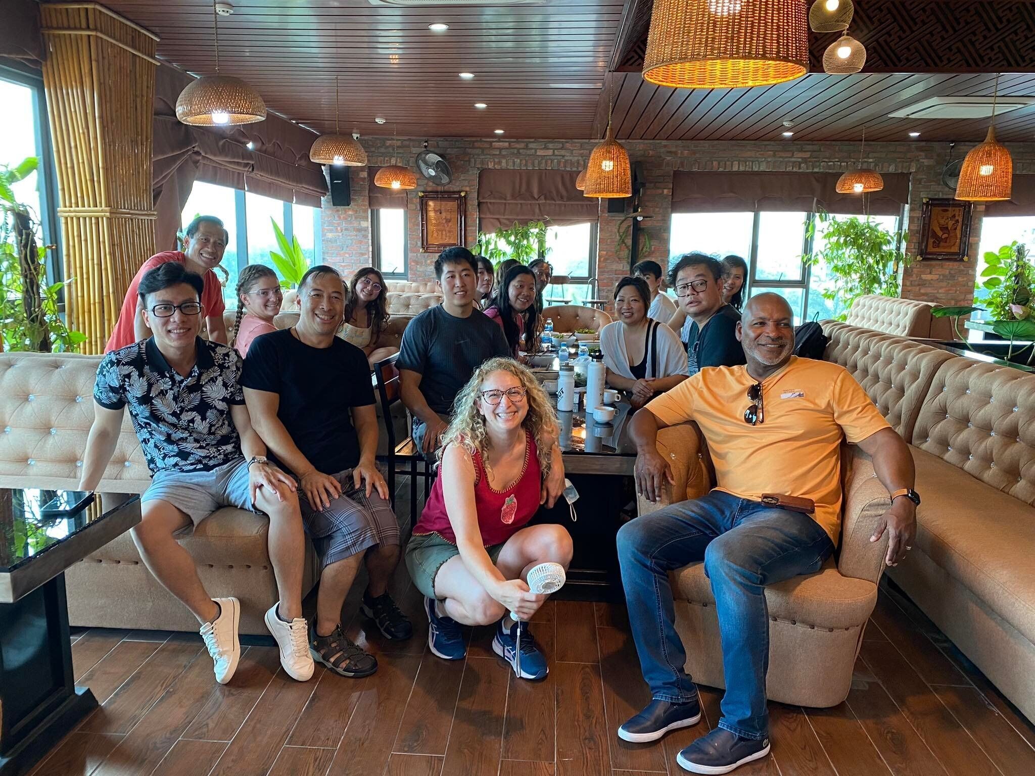 A group of people seated in a restaurant with wooden flooring, brick walls, and hanging wicker lamps.