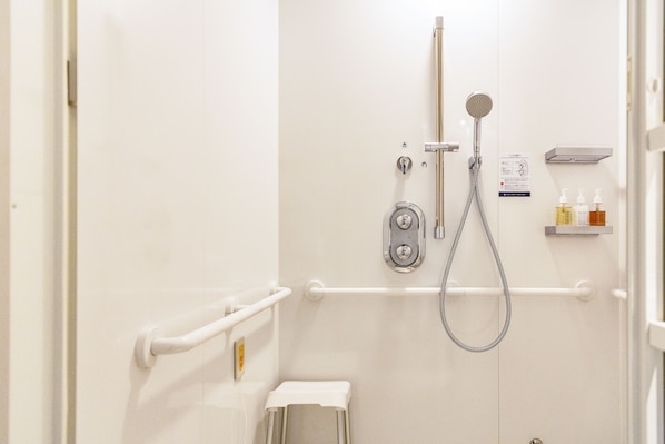 A shower area with a handheld showerhead, a wall-mounted faucet, and a handheld shower hose. There is a white towel on the floor, a small step stool, and a shelf with toiletries. - Hotel Hanshin Annex Osaka (Osaka)