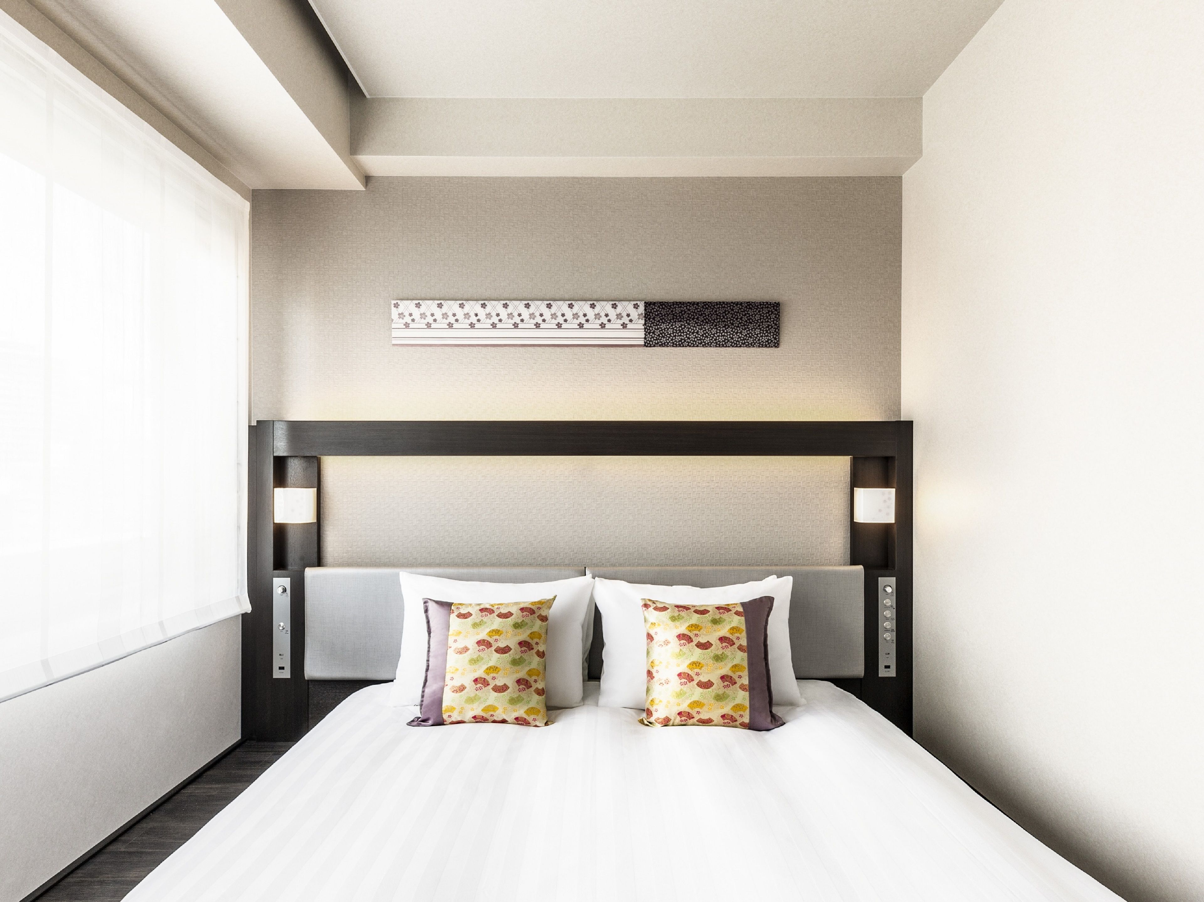 Una habitación de hotel con una cama, dos almohadas, un cabecero, una lámpara fijada a la pared y un panel decorativo sobre la cama.