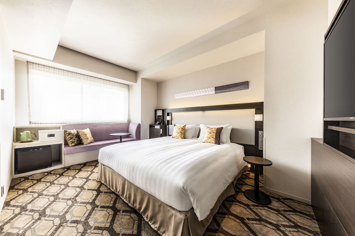A hotel room with a large bed, a seating area with a sofa and pillows, a small table, a coffee machine, and a TV.