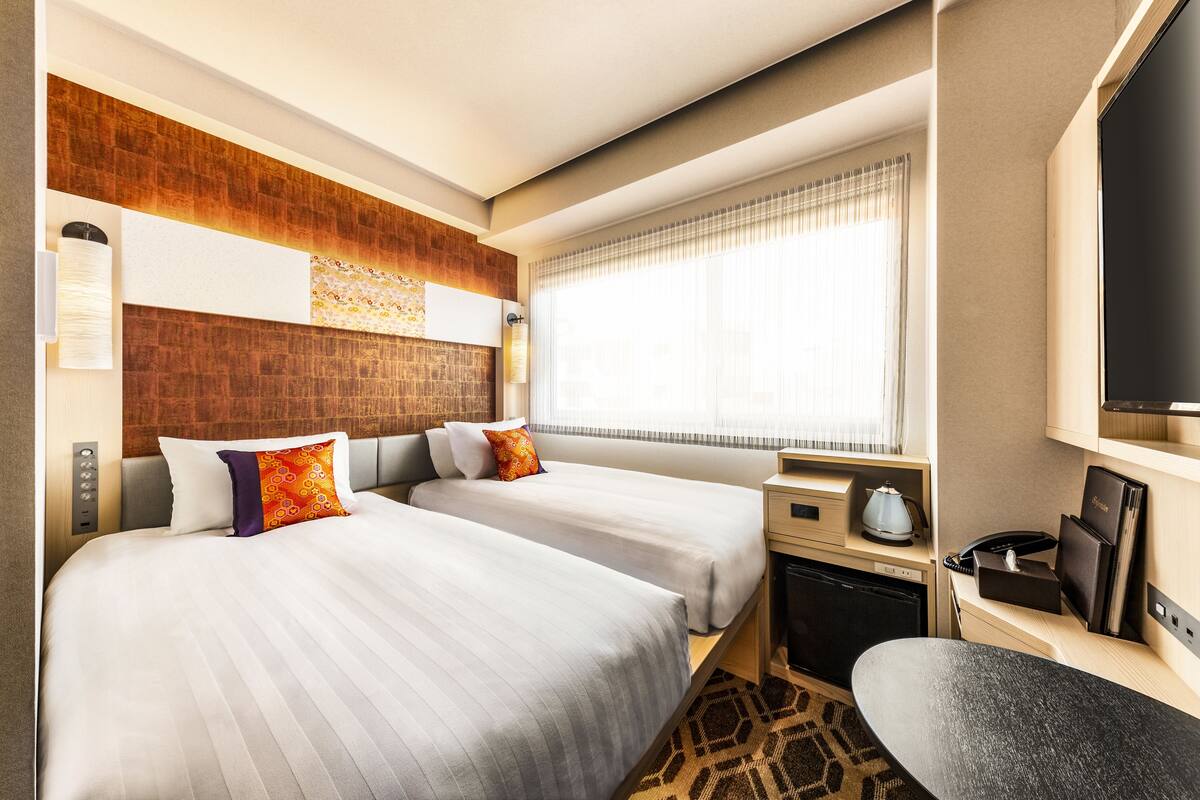 A hotel room with two beds, a desk, a coffee maker, a TV, and a window with curtains.