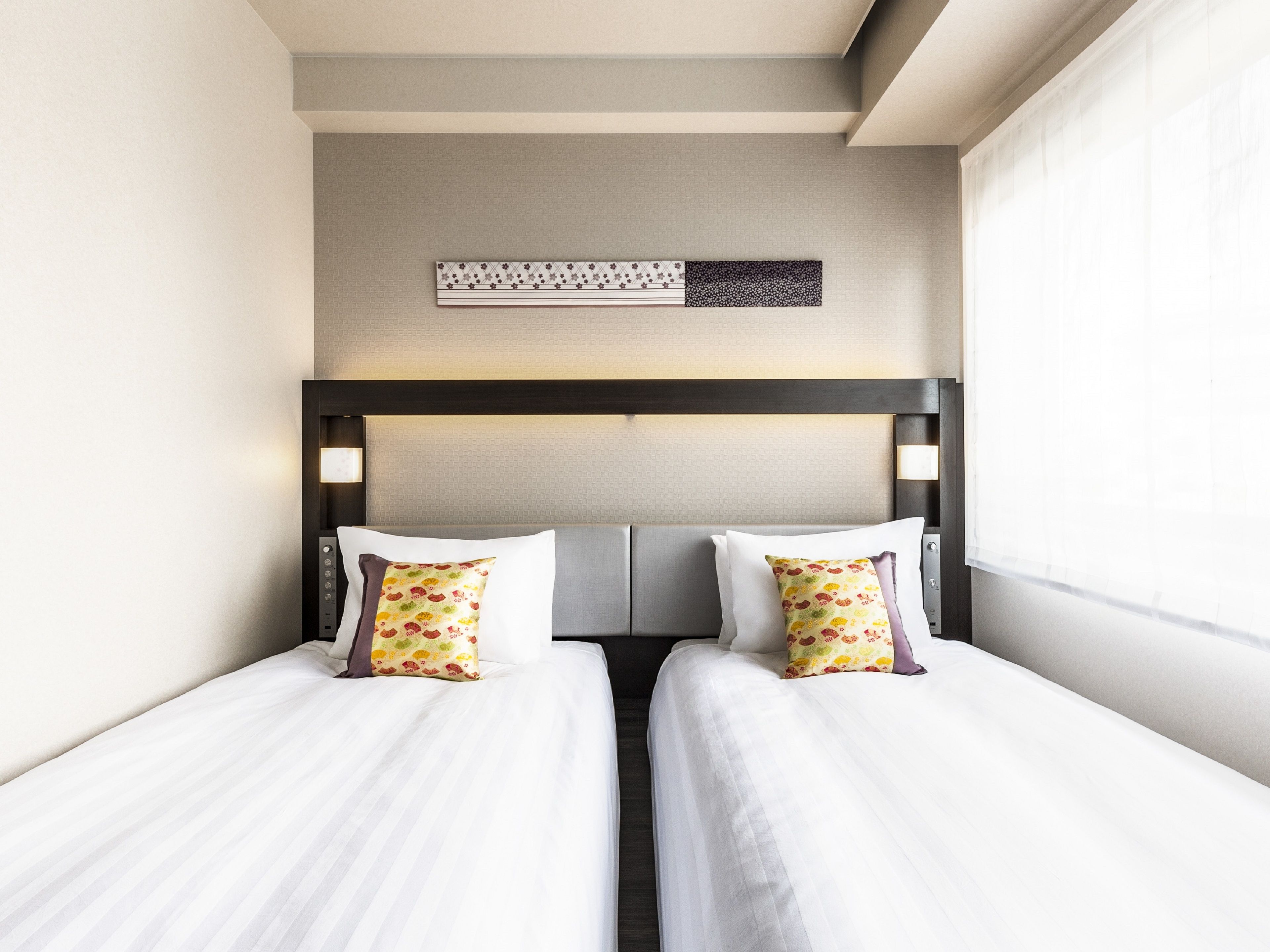 A hotel room with two beds, a headboard with a light fixture, and a decorative wall piece above the beds.