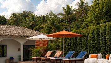 Un área de piscina con sillones de descanso y sombrillas, rodeada de exuberante vegetación y un cielo despejado.