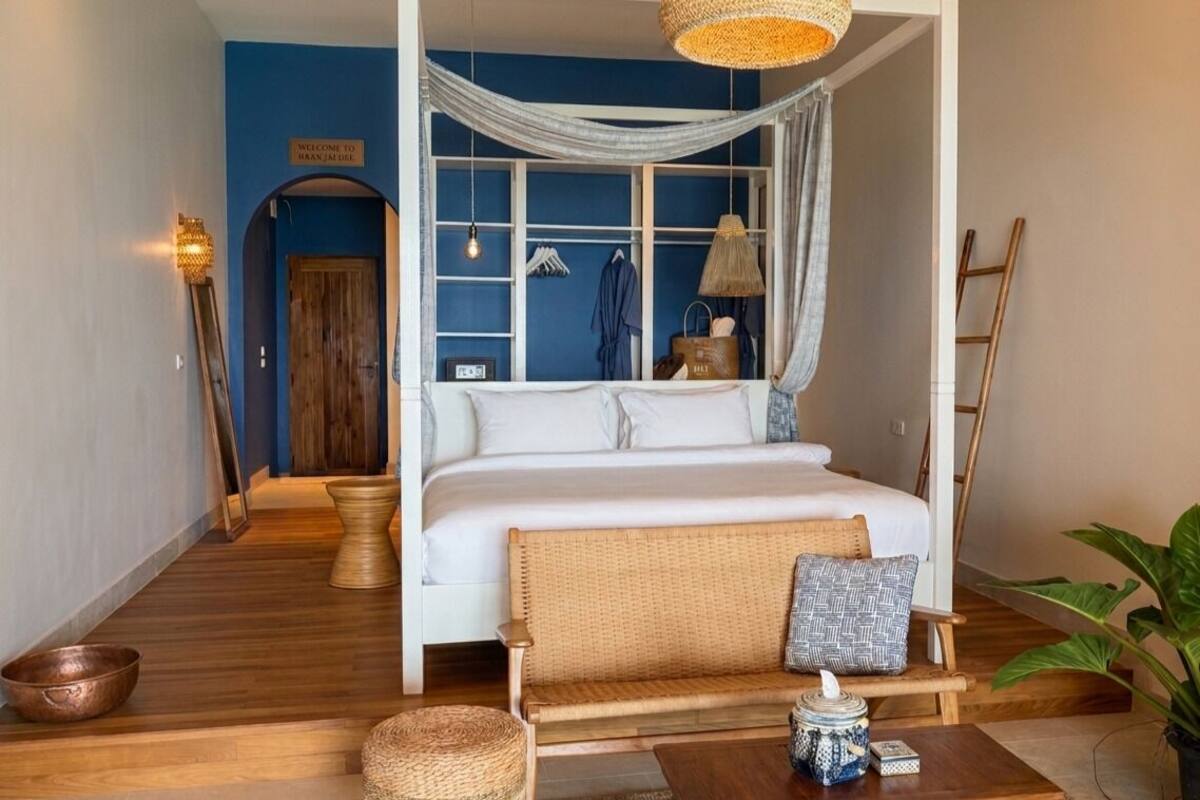 Une chambre à coucher avec un lit à baldaquin, un plancher en bois, un banc en osier et un luminaire suspendu.