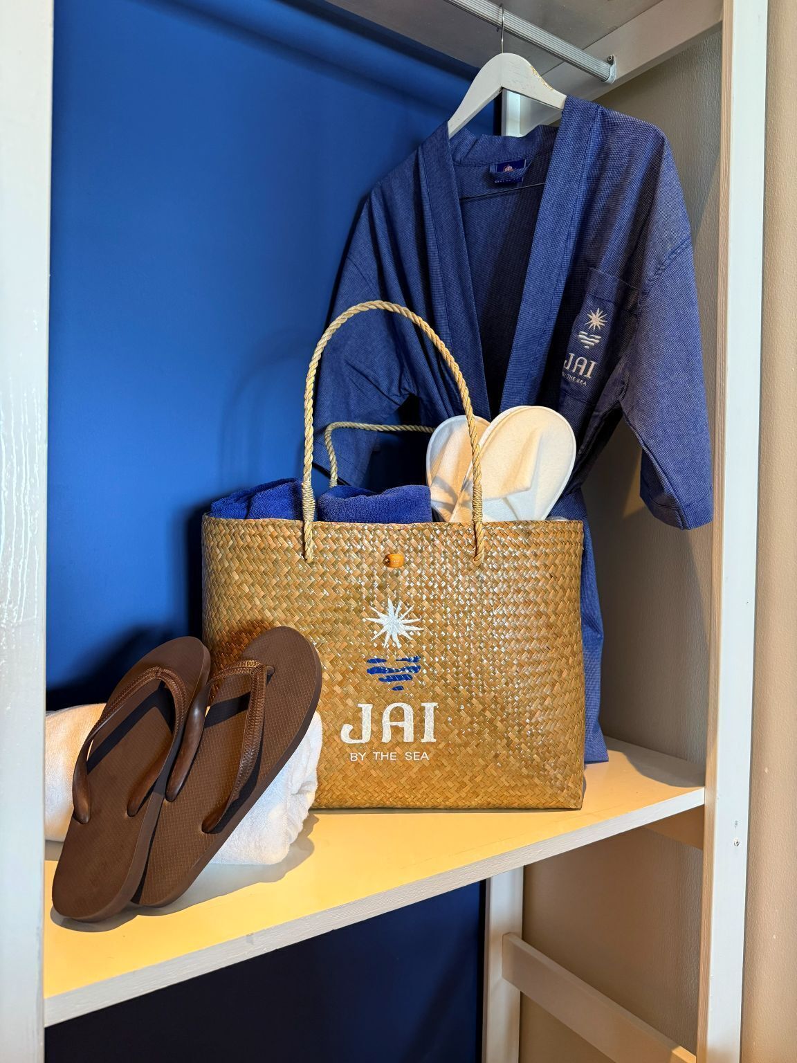 A shelf with a woven bag labeled "JAI BY THE SEA," a pair of brown flip-flops, and a blue garment hanging on a white hanger.