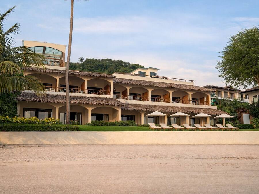 Un hotel de varios pisos con techo de paja, múltiples balcones y una zona de piscina con tumbonas.