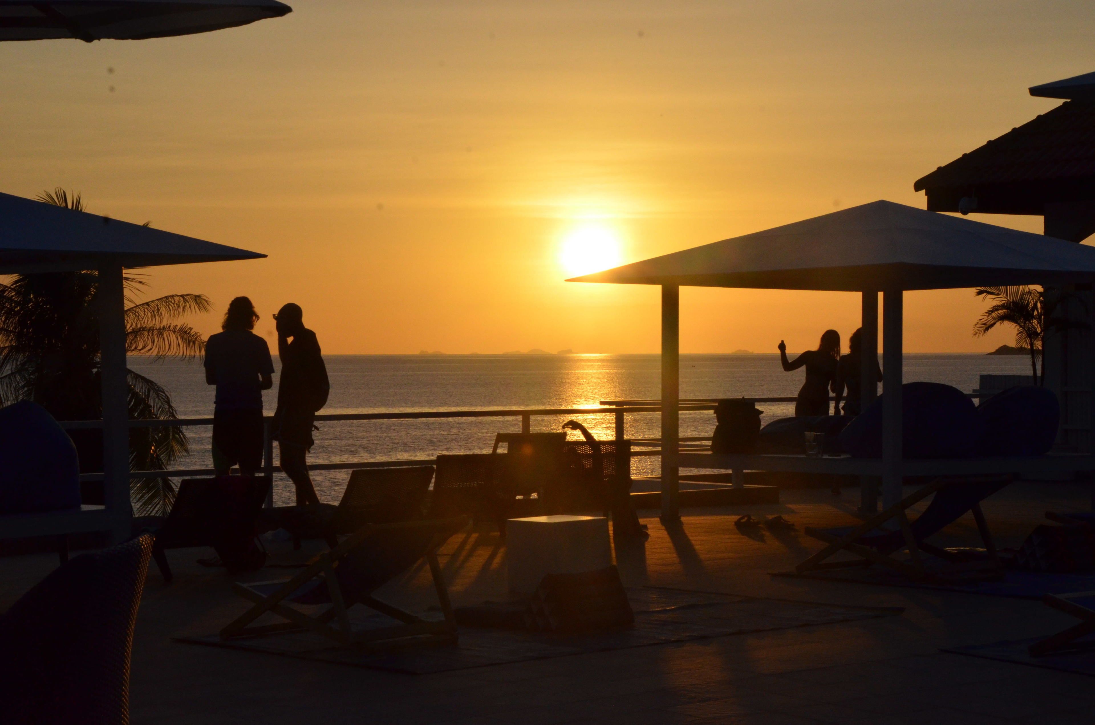 A beachside setting with lounge chairs and umbrellas, a pool area, and a view of the sunset over the ocean.