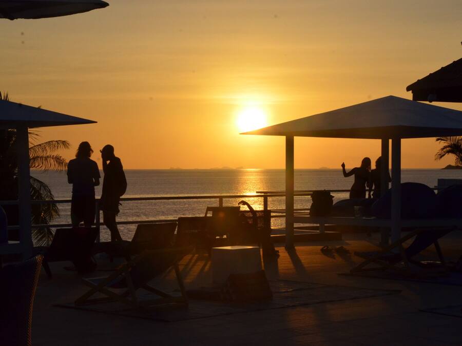 A beachside setting with lounge chairs and umbrellas, a pool area, and a view of the sunset over the ocean.