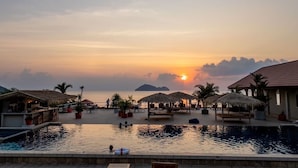 A resort pool area with thatched umbrellas, lounge chairs, and a view of the sunset over the sea. - Baan Jai Dee Beach Front Hotel (Ko Pha-ngan)