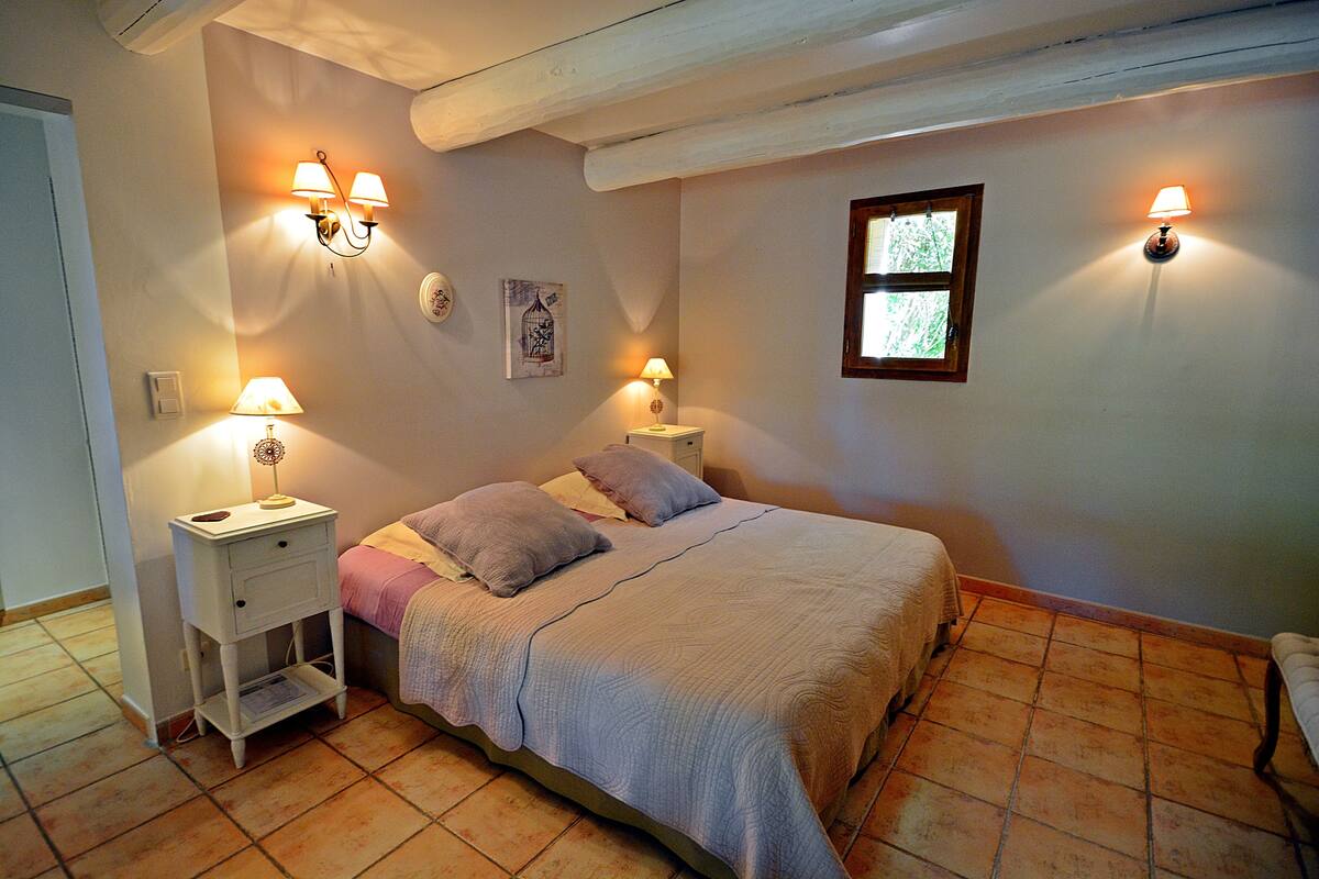 Een slaapkamer met een bed, twee nachtkastjes met lampen, een raam en een wandlamp.