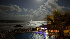 Escena nocturna de una playa con piscina, sillones de descanso y un área de bar.