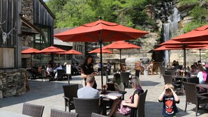 Un área de comedor al aire libre con muros de piedra, sombrillas rojas y una cascada al fondo.
