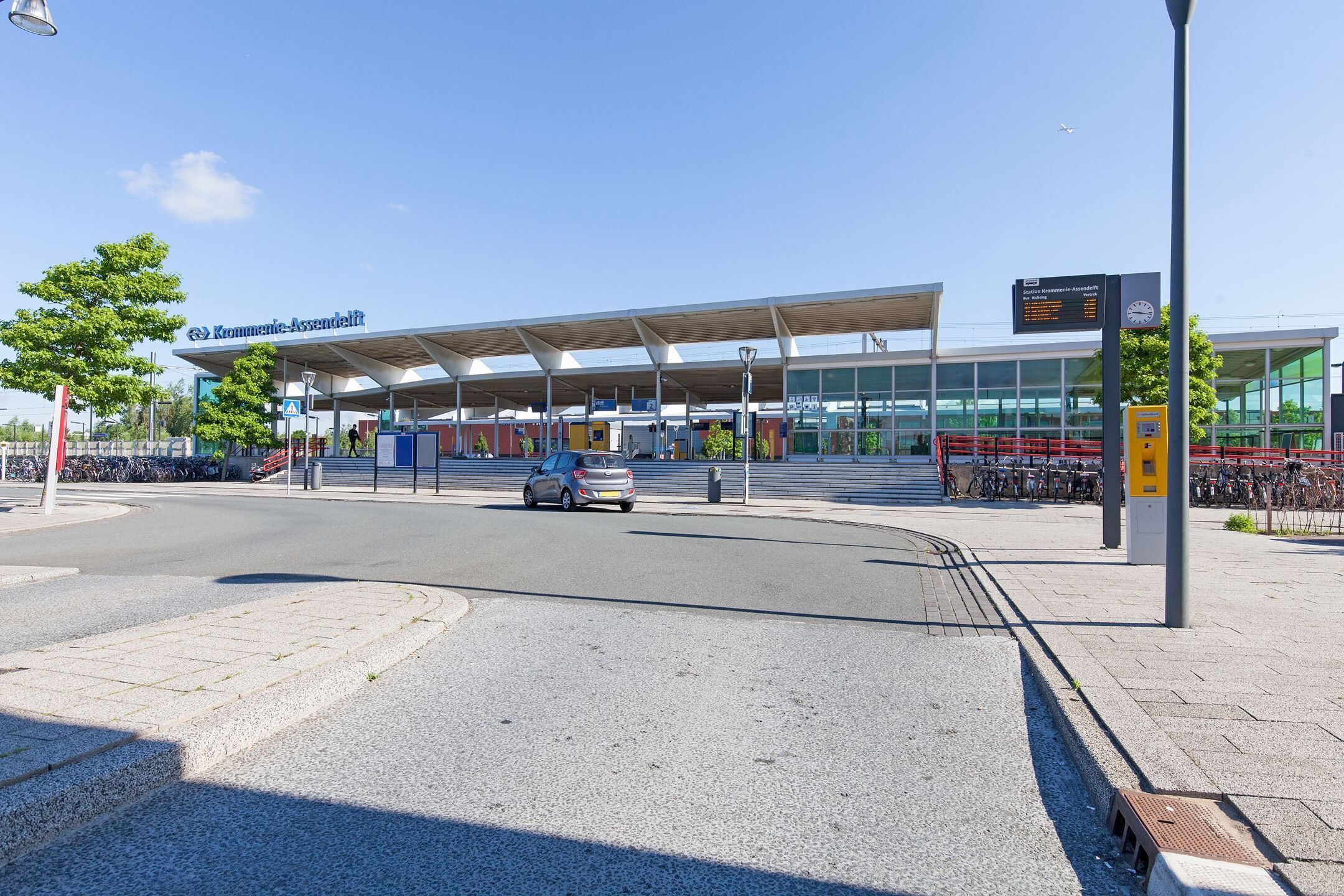 A modern train station with a covered waiting area, a digital information board, and a bicycle parking area.