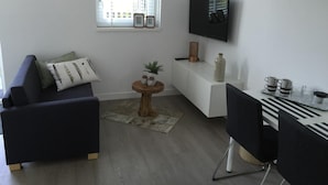 A modern living room with a dark blue sofa, a small round wooden table, a white TV stand, and a dining area with a black chair and a tray of cups and glasses.