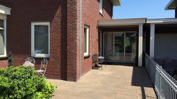 A red brick house with a covered patio, a white door, and a small garden area with a bench and potted plants.