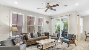 A spacious living room with a ceiling fan, large windows, a sofa set, a coffee table, and a view of the outdoors. - Modern True Blue: Gulf Shores Escape w/ Deeded Beach Access, Pool & Beach Gear! (Gulf Shores)