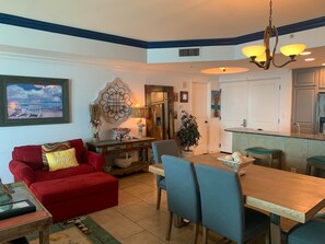 A cozy living room with a red sofa, a wooden dining table, and blue chairs. There is a kitchen area with a white cabinet and a countertop.