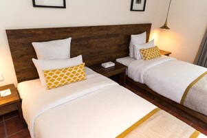 Quarto de hotel com duas camas, cabeceiras de madeira, roupa de cama branca e uma mesa de cabeceira com abajur.