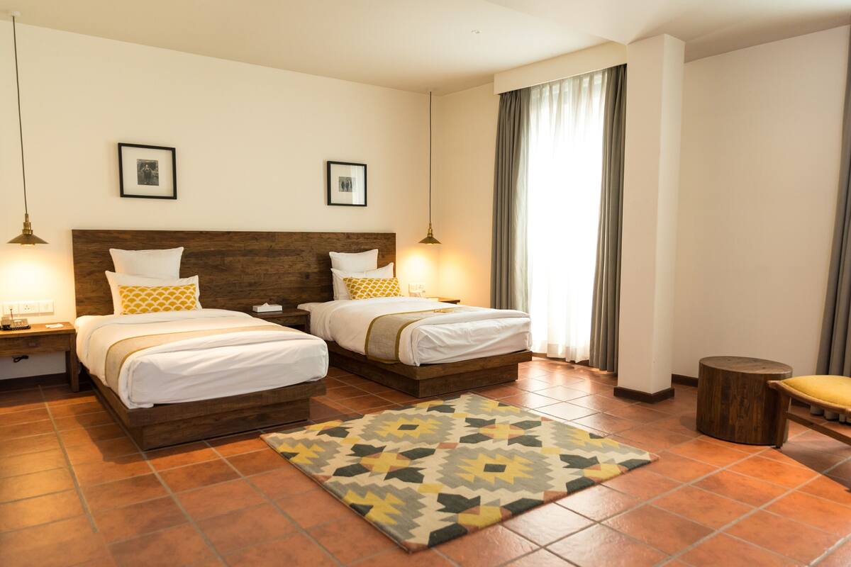 A hotel room with two beds, a wooden headboard, a patterned rug, and a small wooden stool.