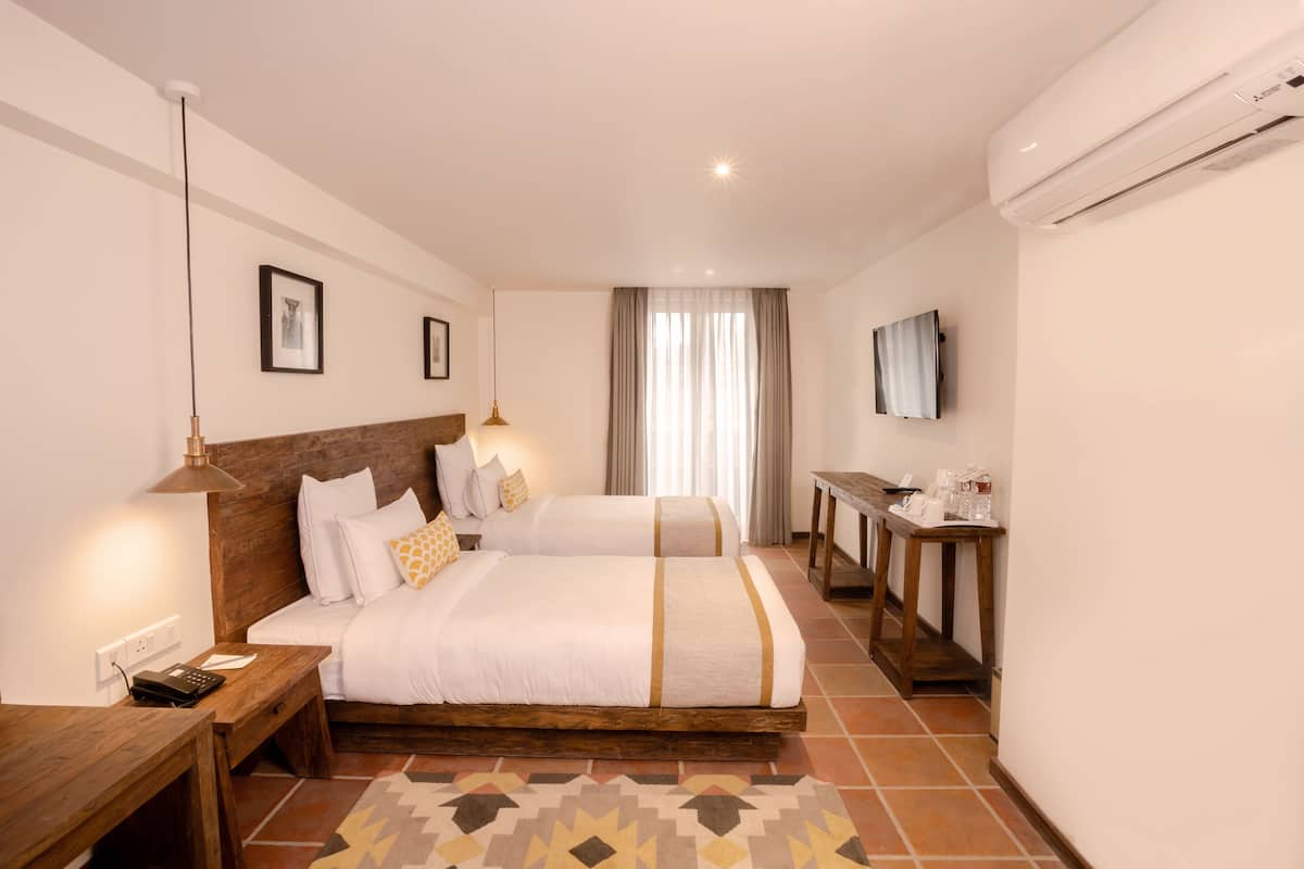 A hotel room with two beds, a desk, a chair, a TV, and an air conditioning unit.