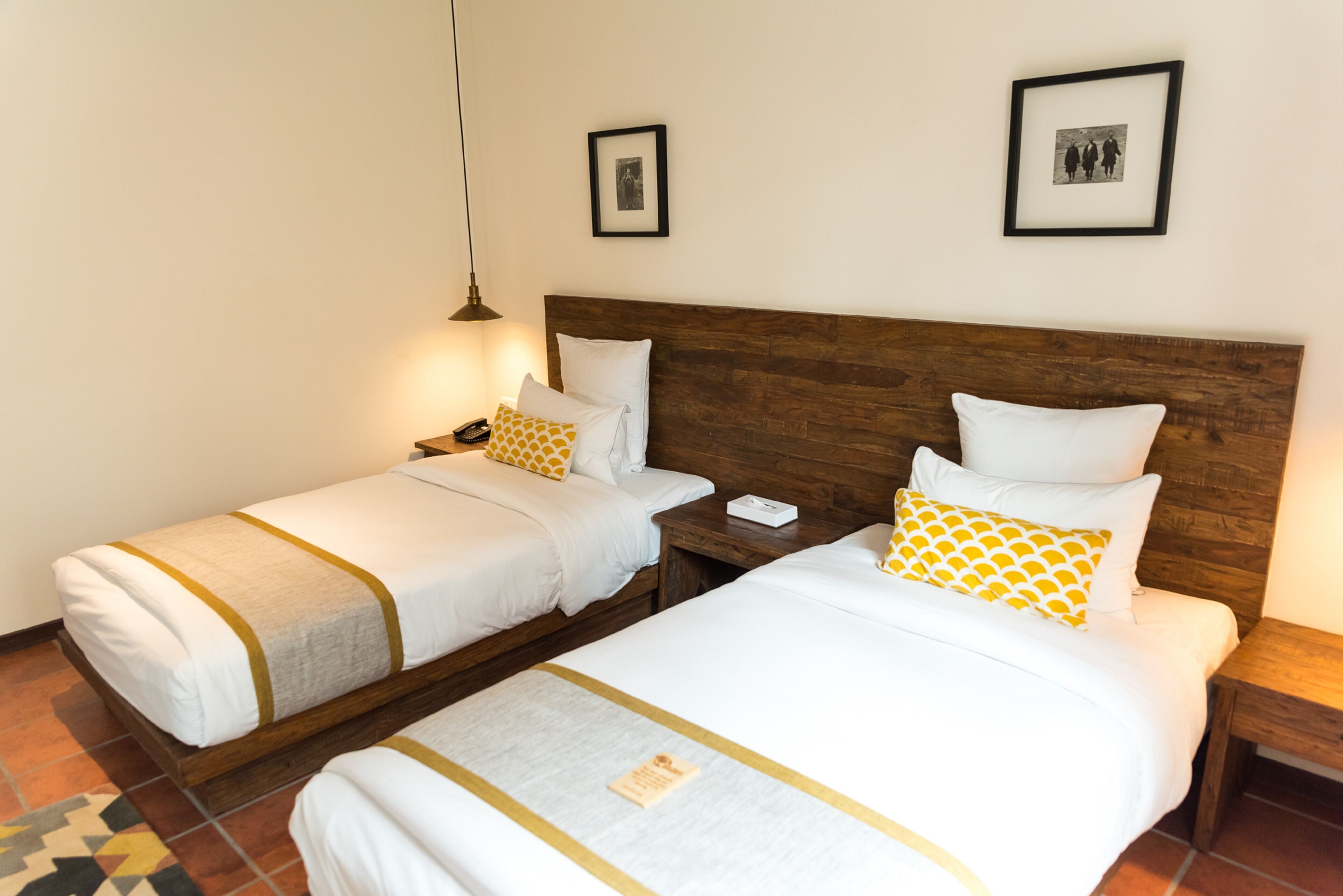 Habitación de hotel con dos camas, cada una con ropa blanca y detalles en amarillo, cabeceros de madera y piso de baldosas.