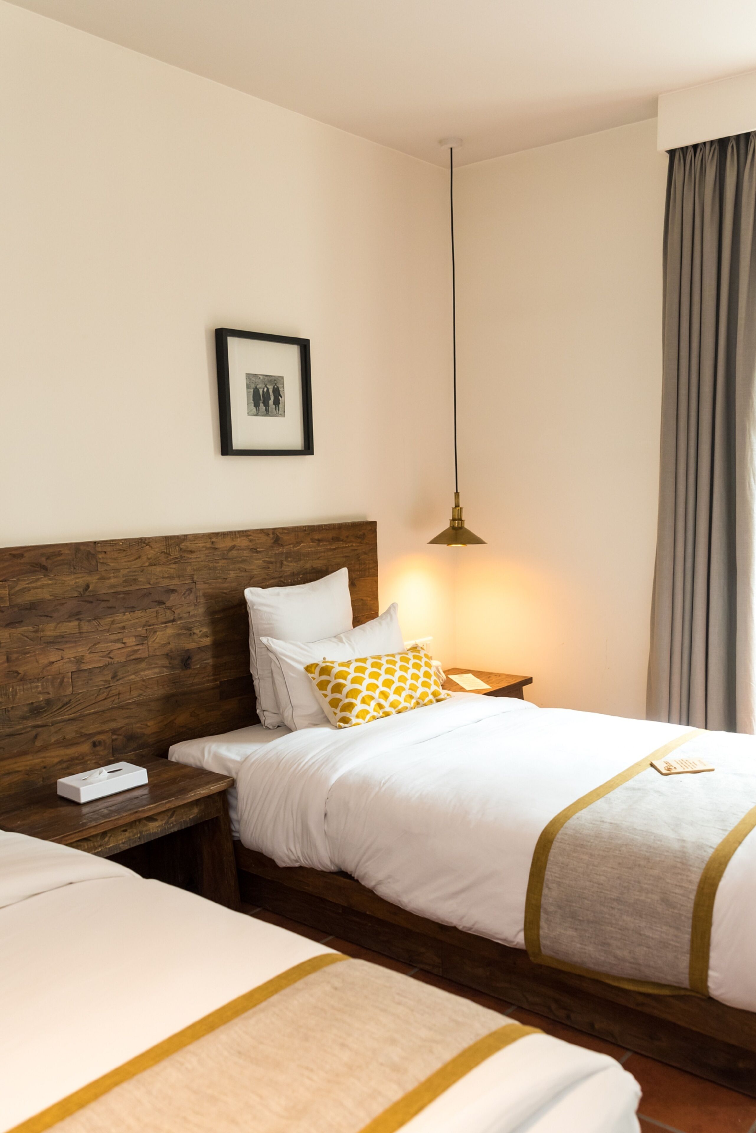 Una habitación de hotel con cabecera de madera, cama con ropa blanca y almohada amarilla, mesita de noche con lámpara, y un cuadro enmarcado en la pared.