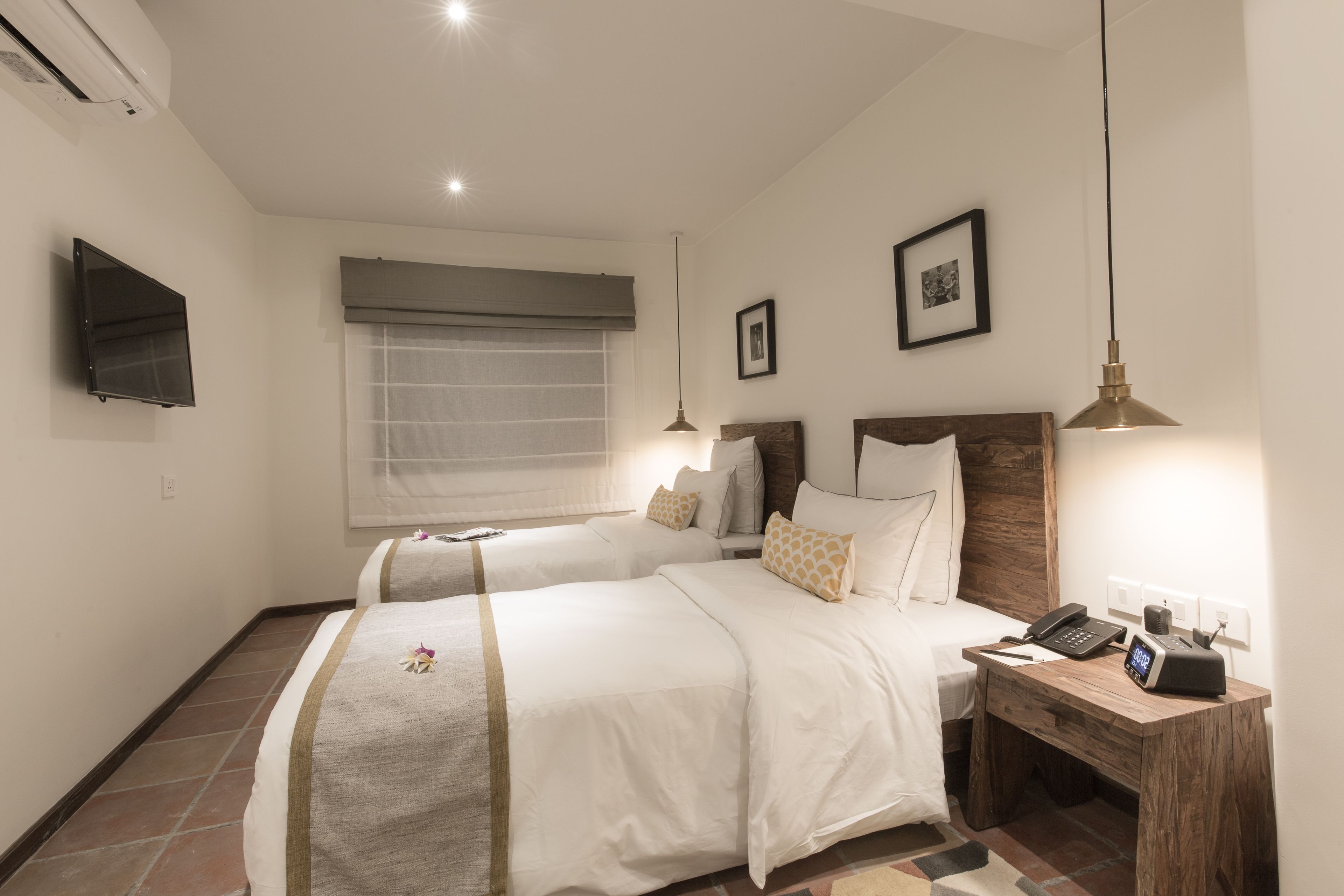 A hotel room with two beds, a bedside table with a phone and a lamp, a TV on the wall, and a window with a curtain.