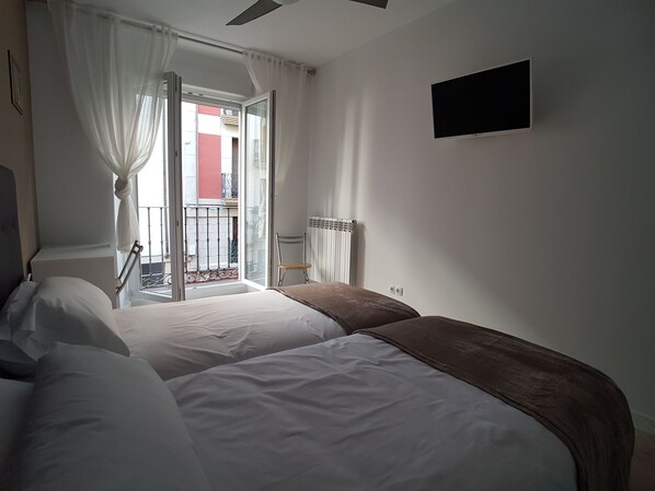 Un dormitorio con dos camas, un radiador, una televisión y un balcón con vistas a edificios.