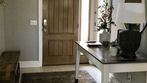Ein Flur mit einer Holztür, einer Bank, einem Tisch mit einer Lampe, einer Vase mit Blumen und einer Chandelier.
