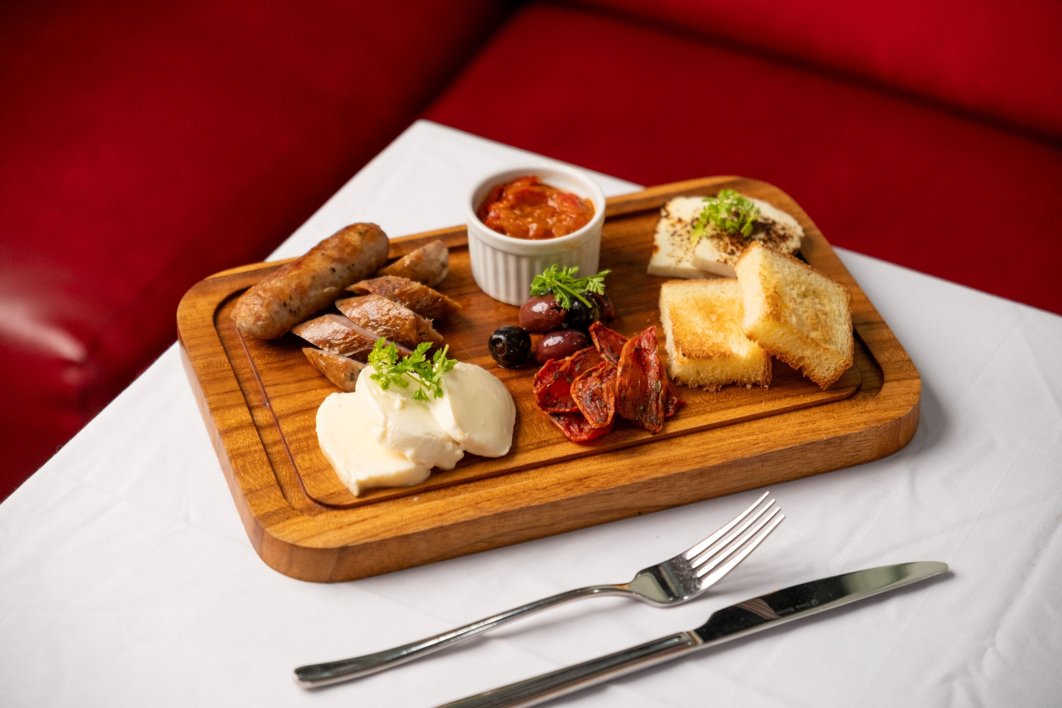 Une planche en bois sur laquelle sont disposés divers aliments, notamment des saucisses, du fromage, des olives et du pain, accompagnée d’un petit bol de sauce, ainsi qu’une fourchette et un couteau, le tout posé sur une nappe blanche.