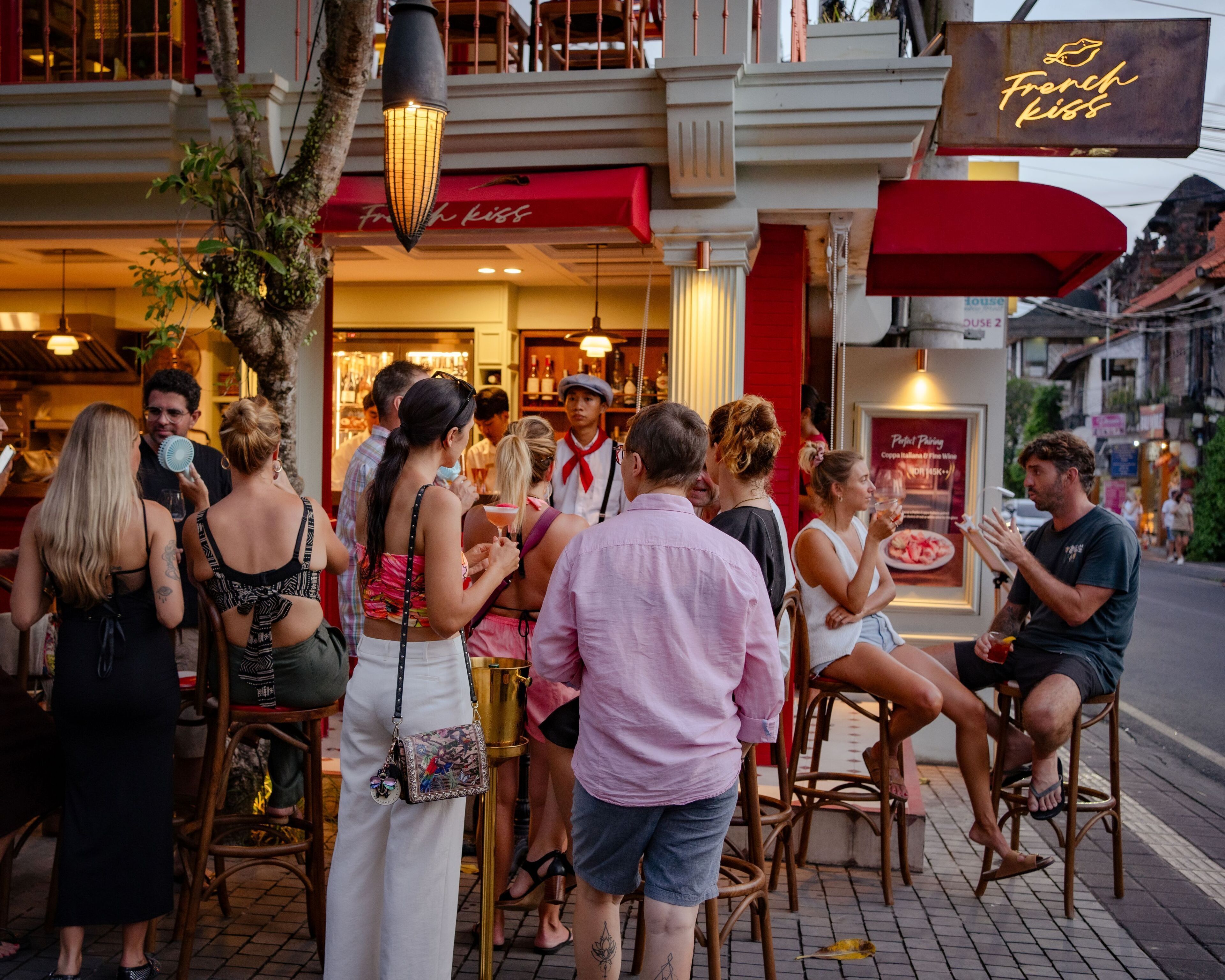 Un grupo de personas reunidas afuera de un restaurante con toldo rojo y un letrero de neón que dice "French Kiss".